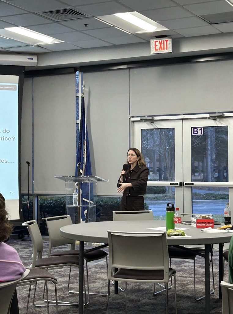 Image of Sarah Silverman presenting. A short white woman with shoulder length brown hair standing and holding a microphone in front of a presentation screen and tables