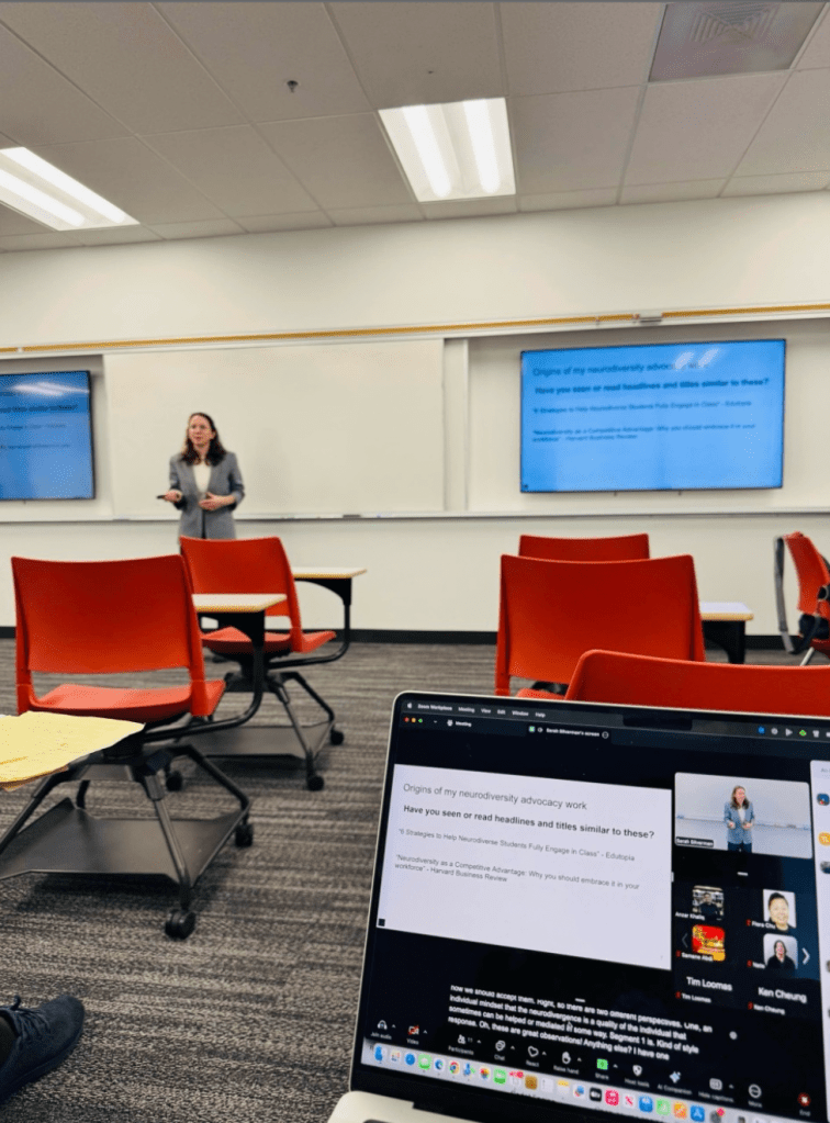 Sarah Silverman (short white woman wearing a blue jacket) standing between two monitors in a classroom with movable desks. A laptop screen connecting the room to remote participants is visible in the foreground